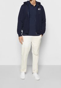 Námořnická mikina na zip s šňůrkami, bílá polokošile a krémové kalhoty. Dokončeno bílými teniskami. Jednoduchý, moderní design, ležérní střih.