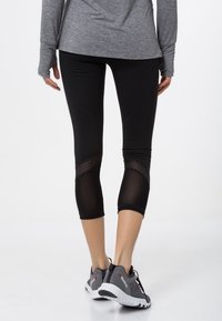 Černé legíny capri s síťovými akcenty a tkaninou odvádějící vlhkost, spárované se šedými sportovními botami s texturovaným svrškem a bílou podešví.