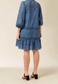Vestido azul de encaje con mangas abullonadas, patrones florales en corte, dobladillo con volantes y cierre de botones en la espalda. Combinado con sandalias negras de tacón cuadrado.