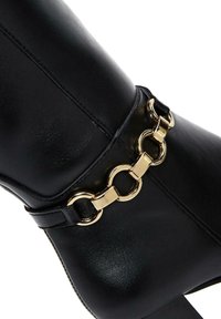 Czarne ankle boots z ekologicznej skóry, zdobione złotym łańcuchem w okolicy kostki, o gładkiej fakturze i ostrej czubek, z wysokim obcasem.