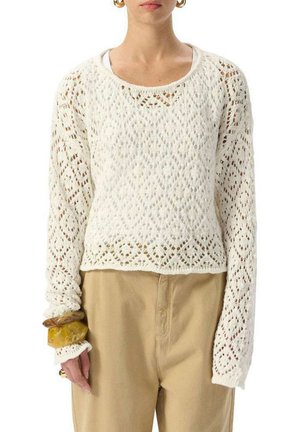 Femme portant un pull blanc à manches longues à mailles ouvertes sur un haut blanc, un pantalon beige et des bracelets épais à son poignet gauche.