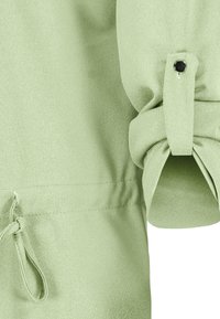 Lichtgroene blouse van een zachte, gestructureerde stof. Voorzien van opgerolde mouwen die met een zwarte knop zijn vastgemaakt en een verstelbare koord in de taille.