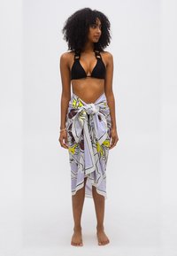 Γυναίκα με σγουρά μαλλιά, που φοράει μαύρο μπικίνι και άσπρο σαρί με κίτρινα και μωβ λουλούδια δεμένο στη μέση, όρθια ξυπόλυτη μπροστά σε απλό φόντο.