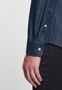 Eine männliche Hand liegt neben dem Körper, die einen dunkelblauen, knöpfbaren Hemdmanschette und schwarze Hosen trägt, vor einem schlichten hellen Hintergrund.