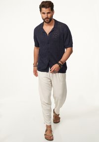 Marineblaues gestricktes Kurzarmhemd mit texturierten Mustern, mit Knopfleiste, getragen mit hellen Leinenhosen und braunen Sandalen.