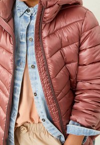 Veste bordeaux matelassée avec une texture brillante, associée à une chemise en jean bleu clair. Un haut beige et un pantalon beige complètent l'ensemble.