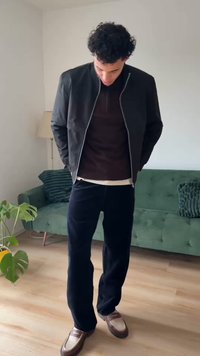 Svart jacka med dragkedja framtill, mörkbrun skjorta och mörka byxor. Bruna loafers med vita detaljer. Grön soffa i bakgrunden.