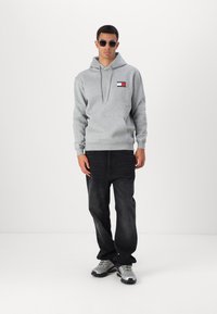 Grå hættetrøje med en lomme foran og et logo-patch, parret med sorte jeans og grå sportsko, iført med mørke solbriller.
