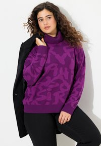 Femme avec de longs cheveux bouclés portant un pull à col roulé à motifs violets et un pantalon noir, tenant une veste noire sur l'épaule devant un fond blanc.