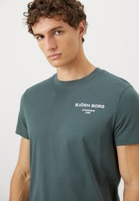 Νεαρός άνδρας με σγουρά μαλλιά, φορώντας ένα πράσινο μπλουζάκι Björn Borg με λευκό λογότυπο και κείμενο "Στοκχόλμη 1956" στο αριστερό στήθος, σε απλό φόντο.