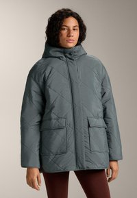 Žena nosi prošivenu, preveliku tamnosivu jaknu s visokim ovratnikom i velikim prednjim džepovima, stoji na neutralnoj pozadini.