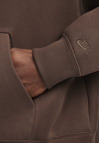 Brun sweatshirt med en glat tekstur. Har en frontlomme og et diskret Nike-logo broderet i en matchende nuance på ærmet.