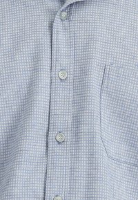 Jasnoniebieska koszula na guziki z delikatnym geometrycznym wzorem, z kieszenią na piersi i szarymi guzikami. Miękki materiał o gładkiej fakturze.