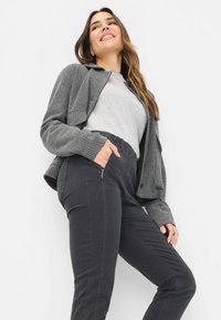 Giacca a spina di pesce grigia, maglione grigio chiaro e pantaloni neri slim-fit con tasche laterali a zip, su uno sfondo bianco uniforme.