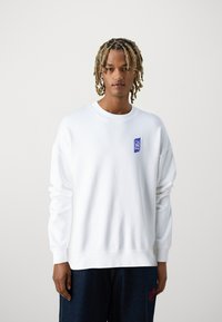 Sudadera blanca con cuello redondo, mangas largas y puños de canalé. Presenta un pequeño parche de logo azul en el pecho. Tejido de algodón suave.