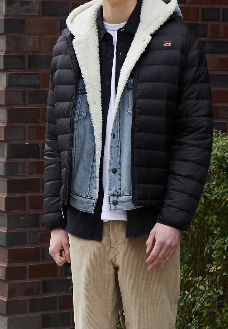 Homme portant des tenues superposées comprenant une doudoune noire avec doublure en polaire blanche, une veste en jean bleu, une chemise noire et un pantalon beige.