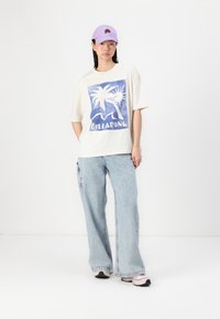 Camiseta gráfica crema de Billabong con estampado de palmeras azules, combinada con unos vaqueros de pierna ancha de un azul claro. Los accesorios incluyen una gorra lavanda y zapatillas.