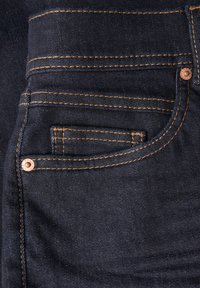 Tejido de mezclilla azul oscuro con una textura suave. Presenta costuras doradas contrastantes, un pequeño bolsillo en la parte delantera y detalles en hardware de cobre.