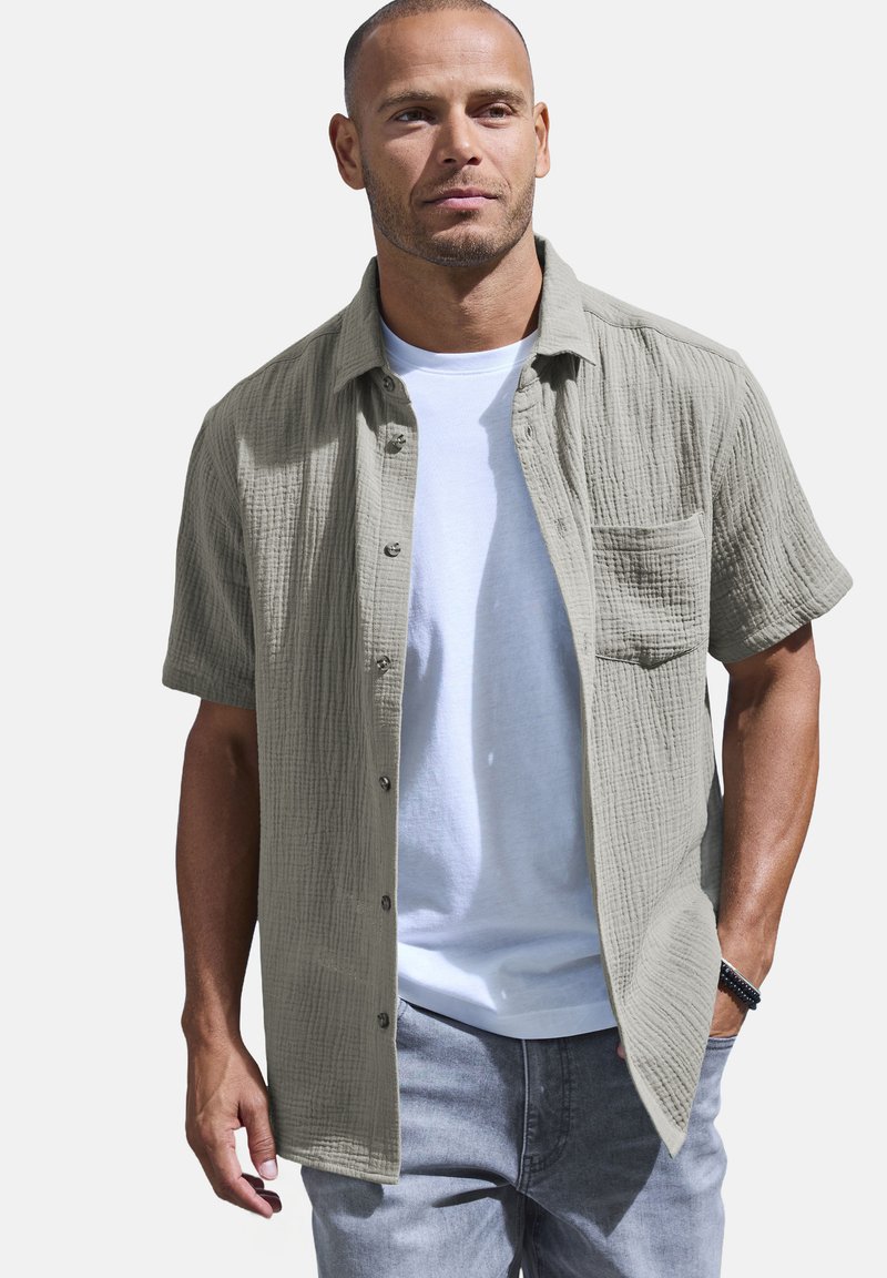 Camicetta grigia chiara a maniche corte con tessuto strutturato, chiusura a bottoni e taschino sul petto, indossata sopra una maglietta bianca e jeans grigi.