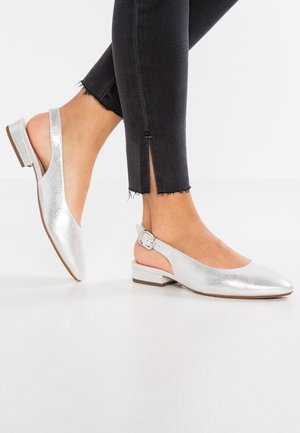 Stopy w srebrnych metalicznych balerinach typu slingback z niskim, blokowym obcasem, w połączeniu z czarnymi, przyciętymi jeansami z bocznym rozcięciem.