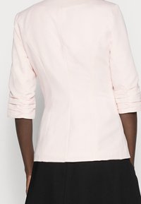 Light pink tailored jacket with three-quarter sleeves, structured seams, and gathered sleeve cuffs, made of smooth fabric.