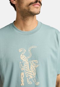 Lichtblauwe katoenen t-shirt met een crèmekleurige graphic van een tijger en het merklogo "RVCA" onderaan. Ronde halslijn, korte mouwen.
