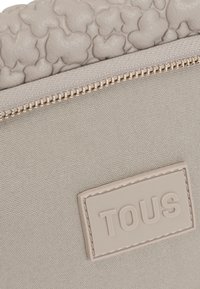 Beżowy portfel z teksturowaną skórzaną górą z kształtami serc, gładkim materiałem na dole, złotym zamkiem błyskawicznym oraz tłoczonym logo "TOUS".