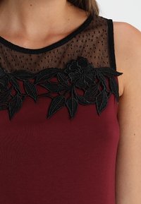 Haut bordeaux sans manches avec un col en tulle noir à pois et une applique florale brodée sur la poitrine.