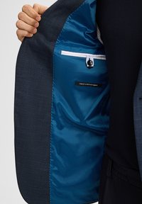 Hand die een open donkerblauwe blazer vasthoudt, met een gladde blauwe voering, binnenzak en label "Finest Stretch Fabric".