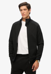 Schwarze Zip-Jacke mit hohem Kragen, aus weichem Stoff gefertigt. Merkmale ein tailliertes Design, kontrastierende Nähte und Seitentaschen. Gestylt über einem weißen Hemd.