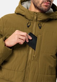 Olivgrön vadderad jacka med dragkedja framtill och en bröstficka. Materialet är texturerat, med en mjuk, fodrad huva.