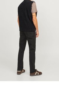 Czarna kamizelka na szarym t-shircie, czarne dżinsy i brązowe sneakersy z jasnymi paskami. Gładka tekstura materiału; luźny krój; minimalistyczny design.