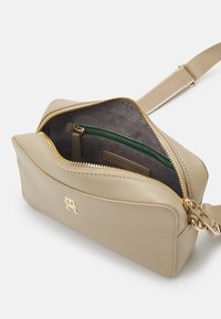 Beżowa skórzana torebka crossbody z złotymi akcentami, zapięciem na zamek oraz zieloną kieszonką zapinaną na zamek w środku. Gładka faktura i wyraźny kształt.