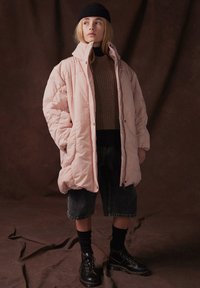 Ljusrosa pufferjacka med hög krage, knappstängning och sidofickor, bärs över en brun stickad tröja och denimshorts, med svarta stövlar.