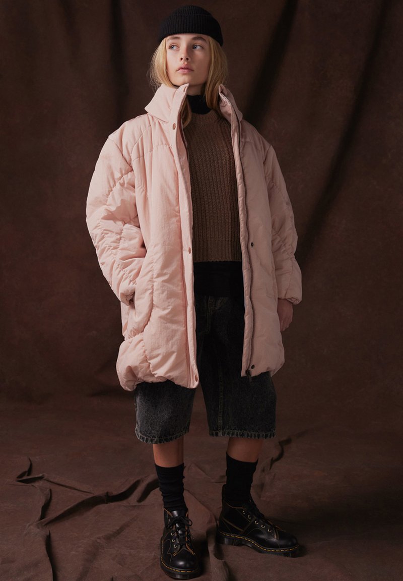 Ljusrosa pufferjacka med hög krage, knappstängning och sidofickor, bärs över en brun stickad tröja och denimshorts, med svarta stövlar.