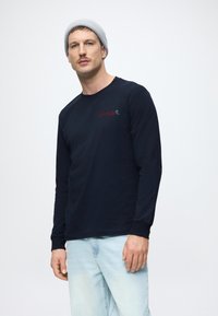 Granatowa koszula z długim rękawem z bawełny, z okrągłym dekoltem i mankietami; zawiera czerwono-niebieskie logo na piersi. Noszona z jasnoniebieskimi dżinsami.