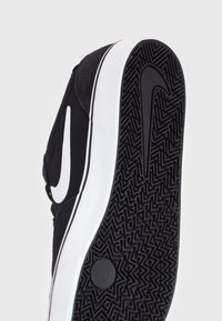 Černo-bílé Nike tenisky ukazující vzorovanou gumovou podrážku s logem Nike swoosh a částí bočního svršku.