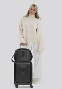Zwart hard-shell bagageset met horizontaal geribbeld ontwerp. De bovenste tas heeft een afneembare schouderriem. Model in lichte trui en witte broek.