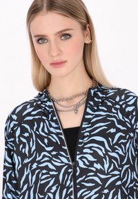 Svart zip-up jacka med ljusblå zebra-randningar. Har en huva, silverdragkedja och lager av kedjehalsband. Texturerat tyg.