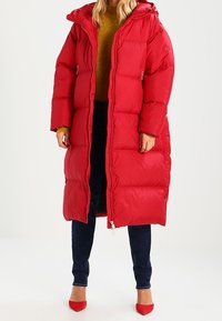 Abrigo largo acolchado rojo con capucha, cierre de cremallera, bolsillos laterales y secciones acolchadas horizontales pronunciadas, combinado con vaqueros oscuros.