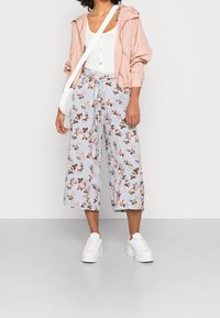 Giacca zip-up rosa chiaro con polsini arricciati sopra un top bianco, abbinata a culotte floreali blu e sneakers platform bianche.
