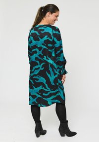 Čierne šaty s tyrkysovým abstraktným vzorom, dlhé rukávy a uvoľnený strih. Nosené s čiernymi pančuchami a členkovými čižmami. Hladká textúra.