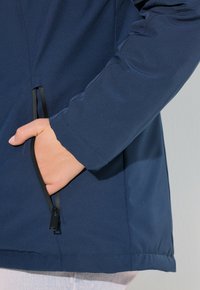 Ruka zasunutá do částečně rozepnuté boční kapsy námořnického modrého saka s dlouhými rukávy, nošeného přes světlé kalhoty.