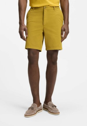 Mand iført sennepsgule shorts og beige ruskind loafers med kvaster, står mod en ensfarvet hvid baggrund.
