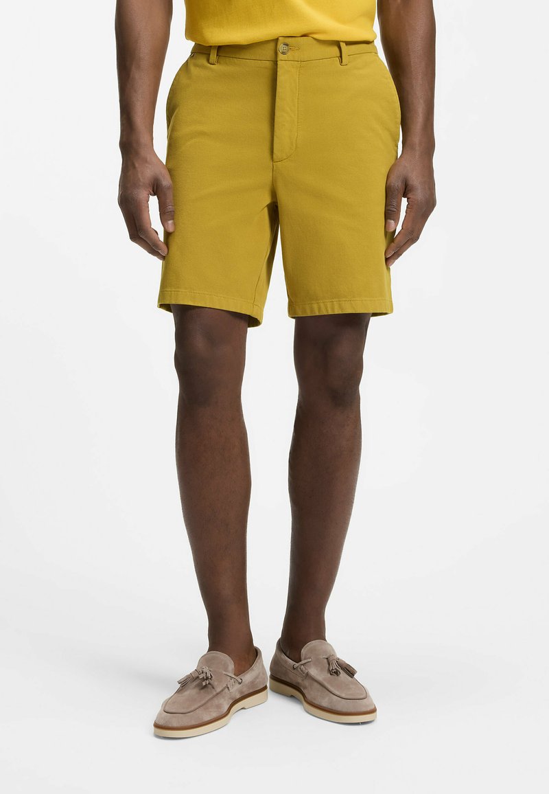 Homme portant un short jaune moutarde et des mocassins à glands en daim beige, debout devant un fond blanc uni.