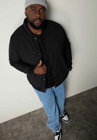 Zwarte button-up gewatteerde jas over een zwart shirt, lichtblauwe baggy jeans en zwart-witte sneakers. Model draagt een lichtgrijze beanie.