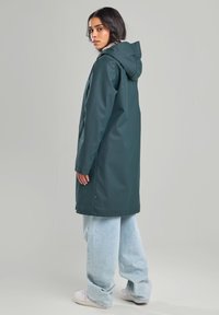 Longue veste imperméable verte avec capuche, dotée de poches latérales et d'une coupe décontractée, portée avec un jean oversize bleu clair et des baskets blanches.