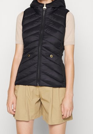 Woman wearing a black quilted sleeveless vest with zipper and gold button pockets over beige short-sleeve top and tan pleated shorts.