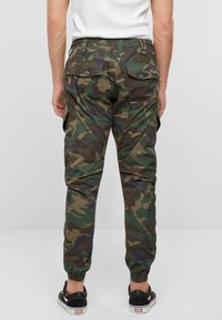 Pantalones cargo en patrón de camuflaje, con cintura elástica, puños ajustados y bolsillos traseros con cierre de botón. El material es una mezcla de algodón.