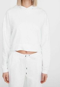 Vit kort hoodie med dragsko och långa ärmar, tillsammans med matchande vita joggers med elastisk midja och svarta detaljer.